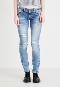 Jean bleu clair déchiré avec une coupe slim, présentant plusieurs déchirures et un design classique à cinq poches. Assorti avec des bottes noires à lacets.