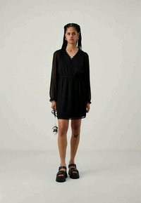 Μαύρο φόρεμα με V-λαιμόκοψη και κυματιστή λεπτομέρεια, μακρυά διάφανα μανίκια με φουσκωτό σχέδιο, σφιχτή μέση και ροή στην φούστα. Φοριέται με χοντρά μαύρα σανδάλια.