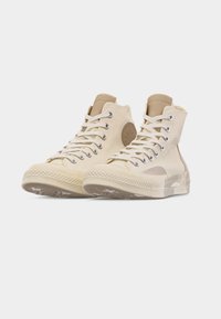 Ζευγάρι μπεζ ψηλά πάνινα παπούτσια με λευκά κορδόνια, λαστιχένιες σόλες και κυκλικό σήμα στον αστράγαλο πάνω σε λευκό φόντο.