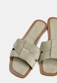 Beige slip-on sandaler med fyrkantig tå, med ett korsremdesign i texturerat material och en platt brun sula.