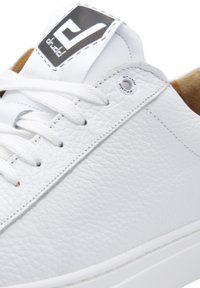 White leather sneaker with textured surface, featuring flat white laces, a logo patch on the tongue, and a tan inner lining.