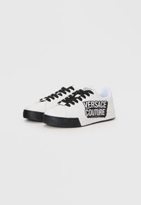 White sneakers with black accents, featuring a black logo on the side, black laces, a textured black sole, and a sleek design.