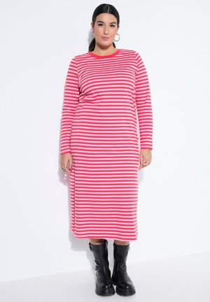 Kvinde med mørkt hår i hestehale iført gyldne hoops, en lang pink og rødstribet kjole og sorte støvler til midt på læggen, stående mod en hvid baggrund.