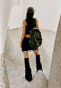 Mochila negra con logo, llevada en un hombro sobre un top corto y una falda negra, combinada con calcetines hasta la rodilla y zapatillas.