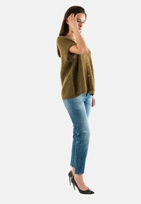Suéter de punto verde oliva con mangas cortas y detalle de botones, combinado con jeans ajustados de mezclilla azul claro y tacones altos negros.