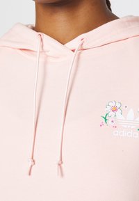Ljusrosa hoodie med dragsko på huvan, prydd med blomstergrafik och adidas-logotyp i vitt. Mjuk textur och avslappnad design.