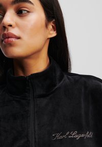 Melna samta džinsjak ar augstu apkakli, kuram krūtīs ir kristālu rotāts "Karl Lagerfeld" logo.