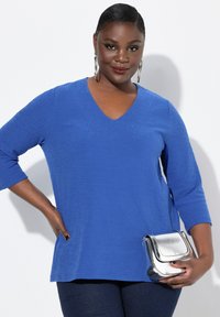 Top azul con escote en V y un patrón geométrico texturizado, de mangas tres cuartos, combinado con un bolso de bandolera metálico plateado.