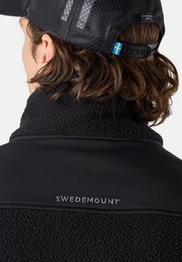 Svart fleecejacka med en texturerad krage, med en slät övre sektion och broderad "SWEDEMOUNT" detalj. Bakifrån syns långt, vågigt hår.