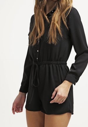 Femme portant une combinaison noire à manches longues avec des boutons sur le devant et une taille nouée, debout devant un fond blanc uni.
