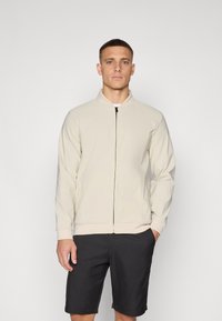 Light beige bomber jacket with a stand collar, front zipper, and ribbed cuffs, paired with black shorts, standing against a neutral background.
