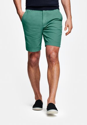 Man draagt groene korte broek, zwarte instappers en een zwart shirt met handen ontspannen langs het lichaam.