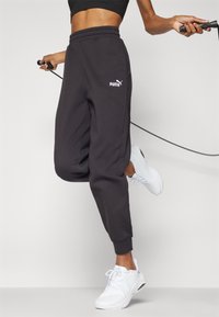 Pantaloni da tuta neri con vita elastica, caratterizzati da un logo Puma bianco, polsini arricciati e vestibilità ampia. Indossati con scarpe sportive bianche.