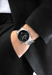 Sudraba nerūsējošā tērauda pulksteņi ar melnu teksturētu ciparnīcu, sudraba stundu rādītāji, datuma funkcija, uz rokas, kas ielikta melnā uzvalka kabatā.