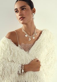 Sølv vedhængsarmbånd med måne- og stjernedesign, sammen med matchende øreringe. Modellen bærer en gennemsigtig beige top og et fluffy hvidt sjal.