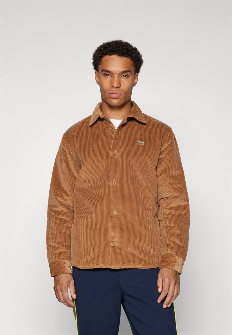 Brown corduroy shirt jacket, buttoned front, collar, ribbed texture, small green Lacoste logo on left chest, fitted design, long sleeves.