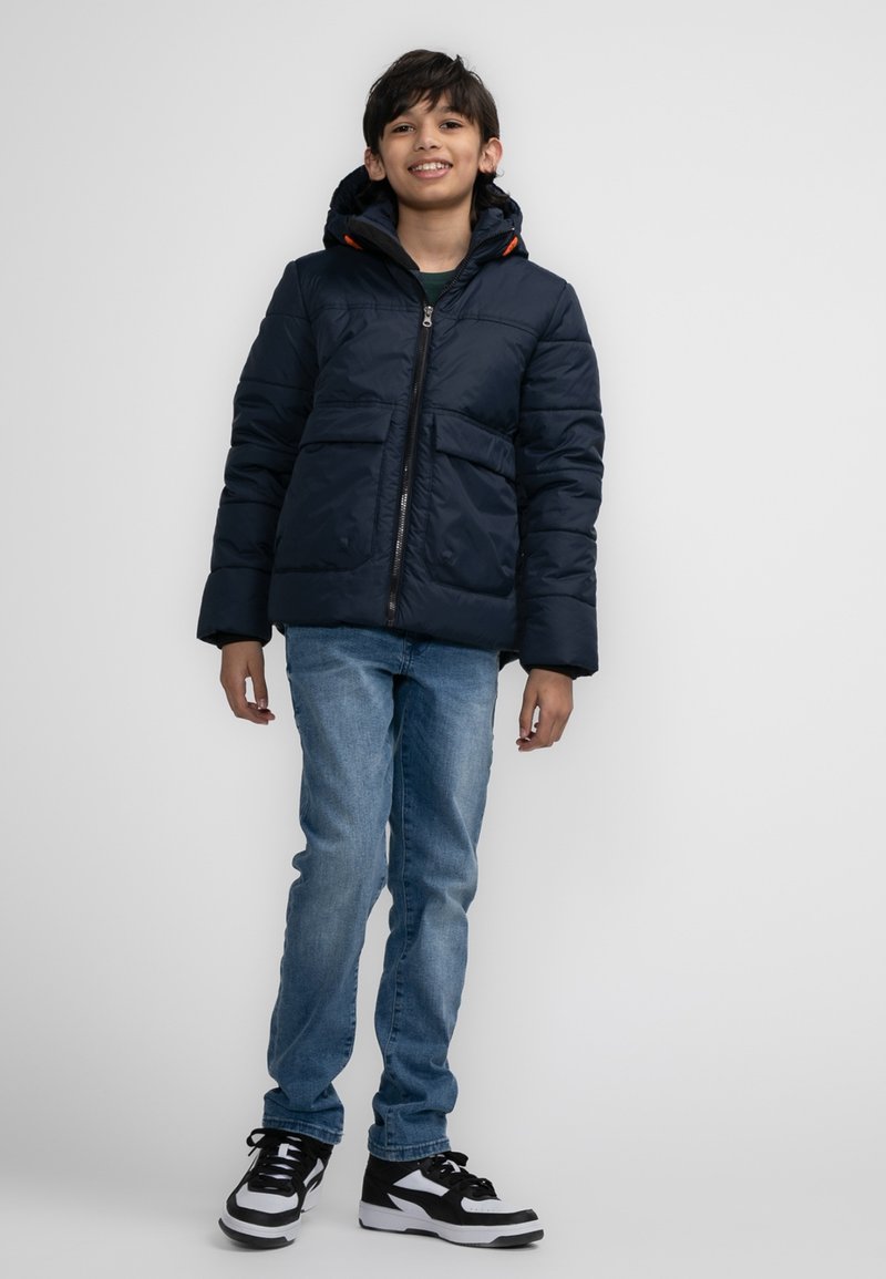 Donkerblauwe gewatteerde jas met capuchon, ritssluiting en twee voorzakken. Gecombineerd met lichtblauwe spijkerbroeken en zwart-witte sneakers.