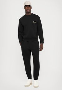 Man wearing black Armani Exchange sweatshirt and pants, white sneakers, and beige Armani Exchange cap, standing against a white background.