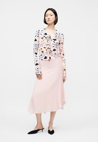 Femme portant un haut à manches longues à fleurs roses, une jupe midi rose clair et des chaussures à talons aiguilles noires, debout devant un fond uni.