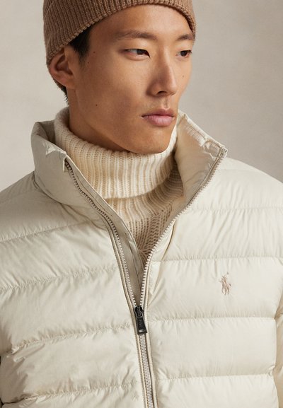 Smelio spalvos pūsta striukė su siuvinėta faktūra ir aukštu apykakliu, dėvima virš kreminės spalvos ribuoto golfuko, su mažyte krūtinės srityje esančia logotipu.