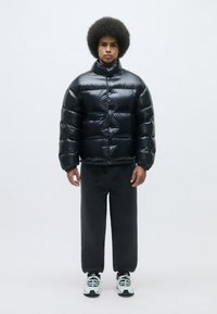 Black puffer jacket with a glossy finish, featuring a high collar, horizontal quilted sections, and a front zip closure. Paired with dark pants and sneakers.
