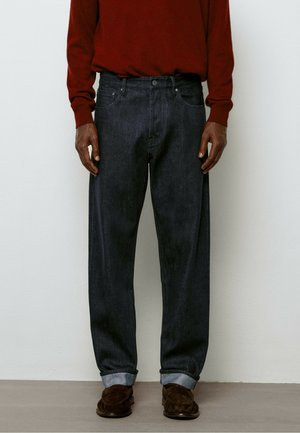 Person iført mørkeblå opkrogede jeans, brune loafers og en rød langærmet sweater står mod en ensfarvet væg.