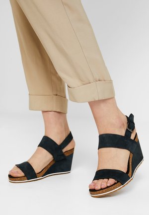 Sandali wedge in suede nero con multiple cinghie, una suola intermedia in sughero e un accento a fibbia. Il bordo bianco della suola contrasta con la calzatura scura.