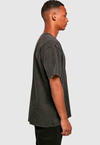 Tricou negru oversized cu decolteu rotund, mâneci scurte și tivuri deteriorate. Prezintă un imprimeu grafic în nuanță de teal pe față.