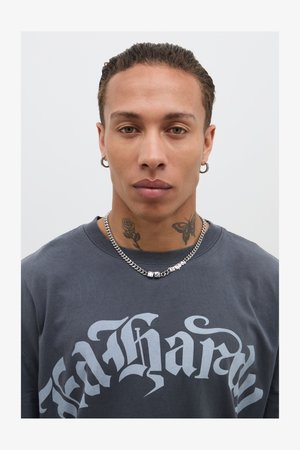 T-shirt grigio scuro con scritte bianche, collana in argento con accenti distintivi e tatuaggi visibili, con un primo piano del viso.