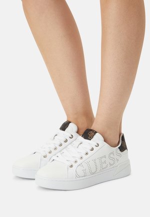 Weiße Sneaker mit niedrigem Schaft, Guess-Logo-Perforation und braunem Muster hinten, getragen an nackten Füßen vor einem schlichten Hintergrund.