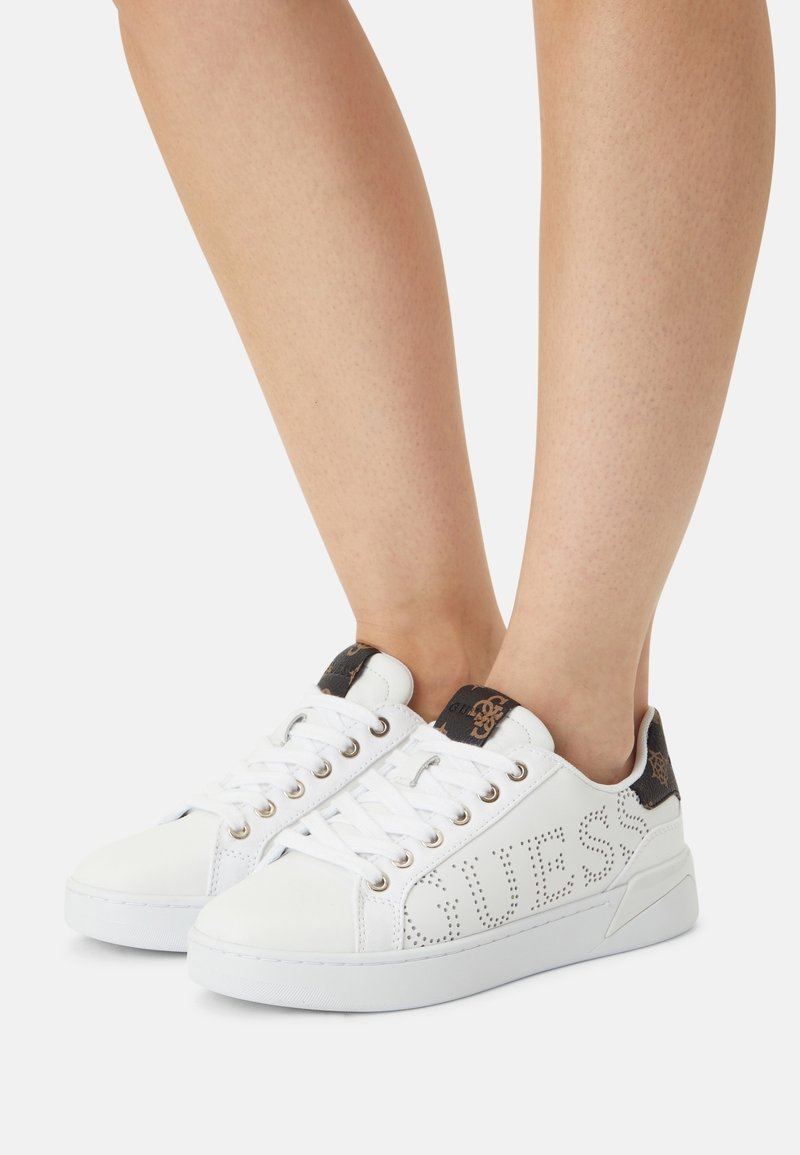 Baskets blanches basses avec perforation du logo Guess et talon marron à motif, portées pieds nus sur un fond uni.