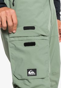Groene waterdichte broek met meerdere zakken, klittenband-sluitingen en een zwart logo-patch op de onderste zak. Glad, functioneel ontwerp.