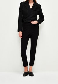 Černý zkrácený blejzr s předními kapsami, spárovaný s černými slim-fit kalhotami a mokasínami. Hladká tkanina a krejčovský design vytvářejí elegantní siluetu.