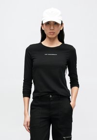 Svart langermet skjorte med hvit merkevare, kombinert med svarte bukser. Modellen har på seg en hvit caps med et opphøyd design.