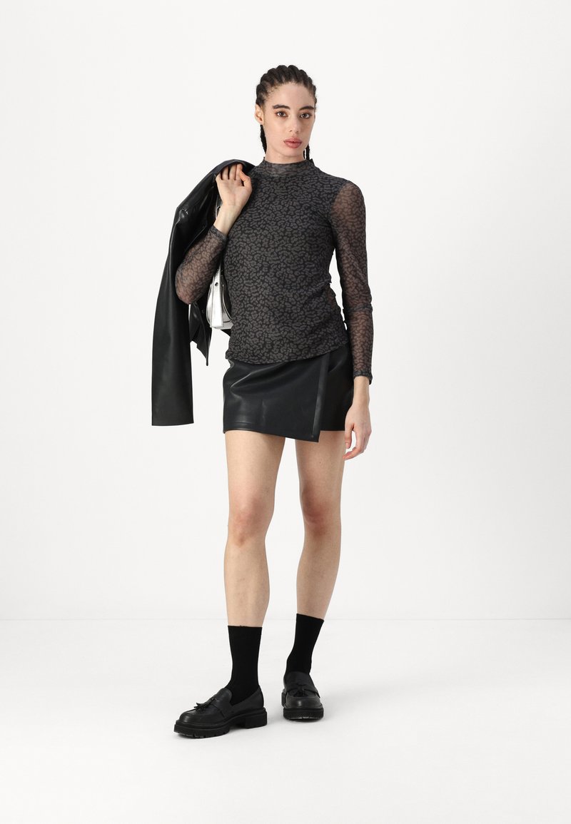 Schwarzes, transparentes Langarmoberteil mit einem subtilen Muster, kombiniert mit einem schwarzen Leder-Minirock. Schwarze Socken und robuste schwarze Schuhe runden den Look ab.