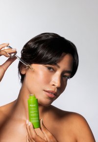 Grön flaska med vit text, utrustad med pipett. Personen applicerar olja på sin kind, vilket visar på jämn hy och mörkt hår.