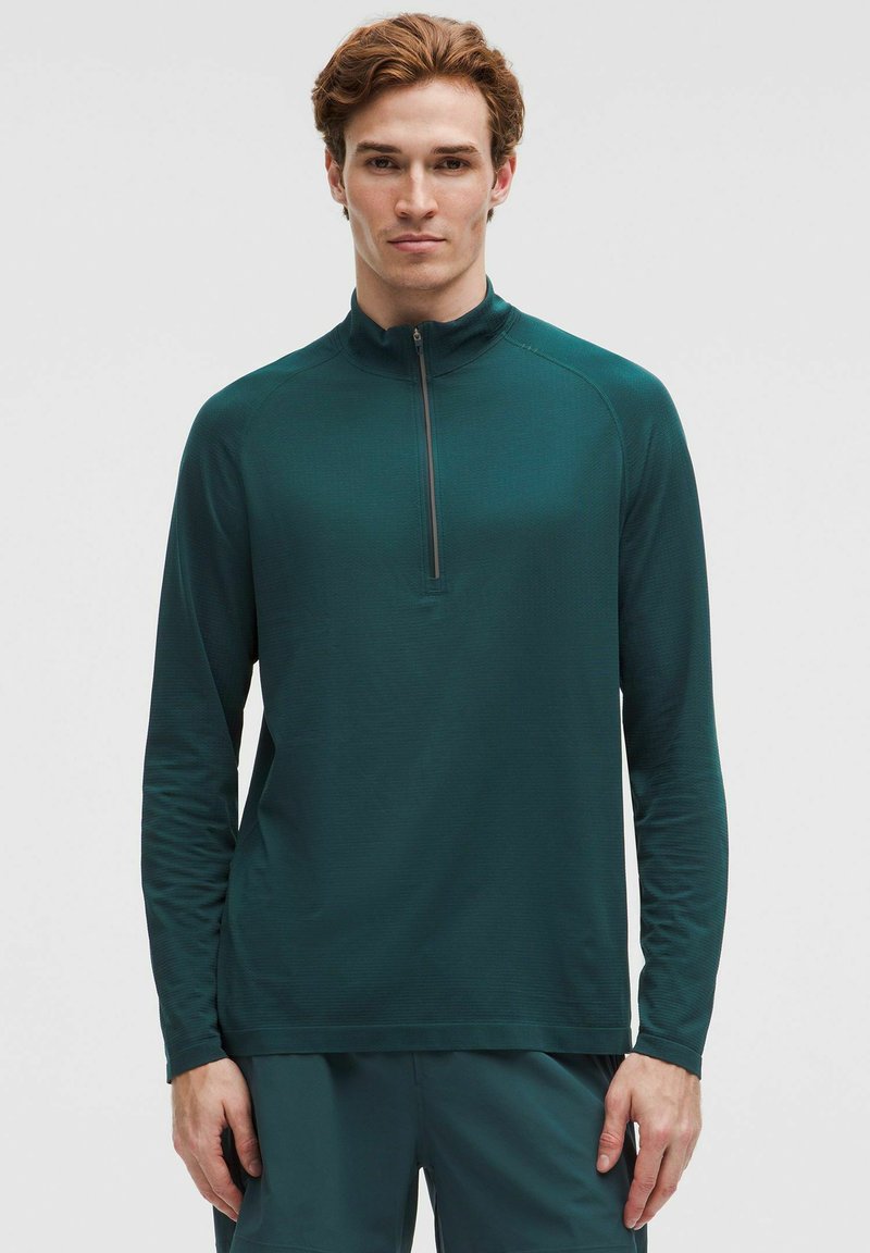 Šviesios odos vyriškis su trumpomis garbanotomis kaštoninėmis plaukų šukuosenomis, vilkintis tamsiai žalią ilgomis rankovėmis polo megztinį su ketvirtinio užtrauktuko uždarymu ir atitinkamas kelnes, stovi atsipalaidavęs.