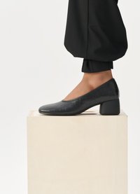 Pantof cu toc din piele neagră, cu un model texturat de crocodil, vârf rotund și toc pătrat, expus pe un piedestal din lemn deschis.