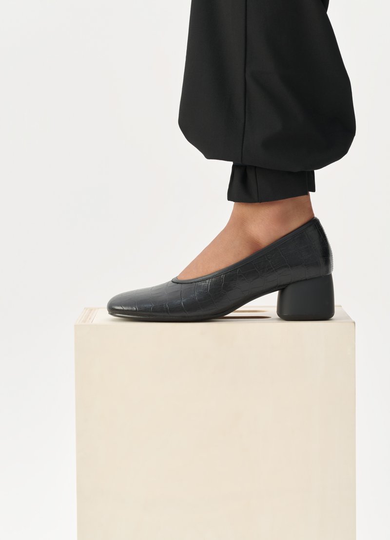 Schwarze Lederpumps mit einer strukturierten Krokodilprägung, runder Zehenpartie und Blockabsatz, auf einem hellen Holzpult präsentiert.