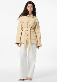 Veste en denim beige clair avec une taille ceinturée, deux poches avant et des fermetures à boutons, associée à un pantalon large blanc et des sandales.