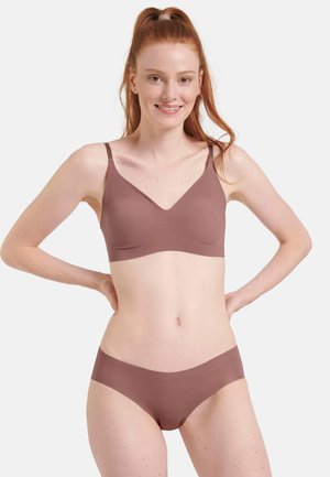 Woman with red hair wearing a mauve seamless bra and matching underwear, standing with hands on hips against a plain background.