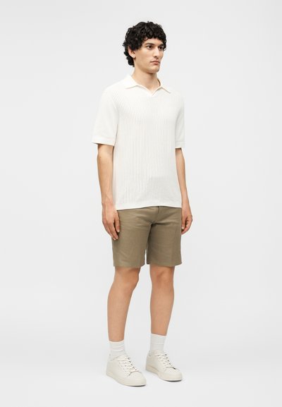 Άνδρας με σγουρά μαλλιά που φοράει λευκό κοντομάνικο πλεκτό πόλο, μπεζ βερμούδα μέχρι το γόνατο, λευκές κάλτσες και λευκά αθλητικά παπούτσια, στέκεται μπροστά σε μια μονόχρωμη επιφάνεια.