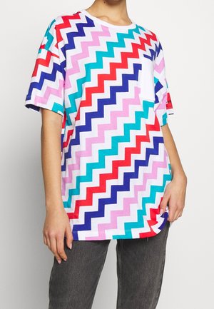 Persoon draagt een wit T-shirt met multicolor zigzagstrepen in rood, blauw, roze en teal, gecombineerd met donkergrijze broek.