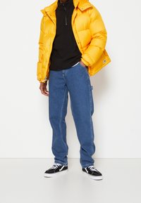 Persona che indossa un giubbotto di piuma giallo brillante, una felpa nera con zip, jeans blu larghi e sneakers in bianco e nero, in piedi su uno sfondo bianco.
