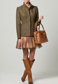 Mujer vistiendo una chaqueta ajustada verde oliva, falda multicolor, botas de cuero marrón hasta la rodilla, sosteniendo un bolso de cuero marrón.