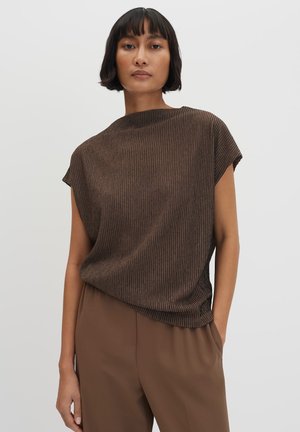 Vrouw met kort zwart haar, gekleed in een bruin geribd mouwloos topje en een bruine broek, staand met één hand in de zak tegen een effen achtergrond.