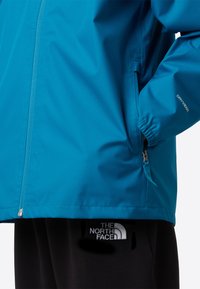 Jachetă impermeabilă albastră, cu fermoar frontal, manșete elastice și buzunar lateral; asortată cu pantaloni negri ce poartă un logo.