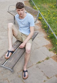 Neformální outfit s světle modrým tričkem, béžovými šortkami a černými žabkami s hnědými podrážkami, sedící na dřevěné lavici v parku.
