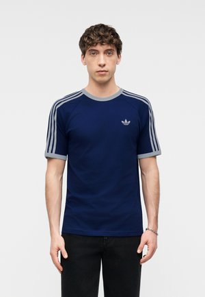 Νεαρός άνδρας με σγουρά μαλλιά, φορώντας μπλε-ναυτικό μπλουζάκι Adidas με γκρι ρίγες στα μανίκια και λογότυπο στο στήθος, στέκεται μπροστά σε απλό φόντο.