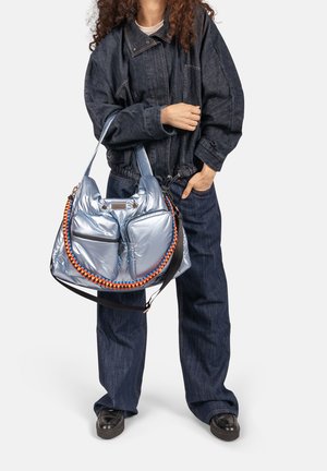 Persoon in donkerblauw denim outfit die een grote glanzende zilveren handtas vasthoudt met een oranje en blauwe gevlochten band.